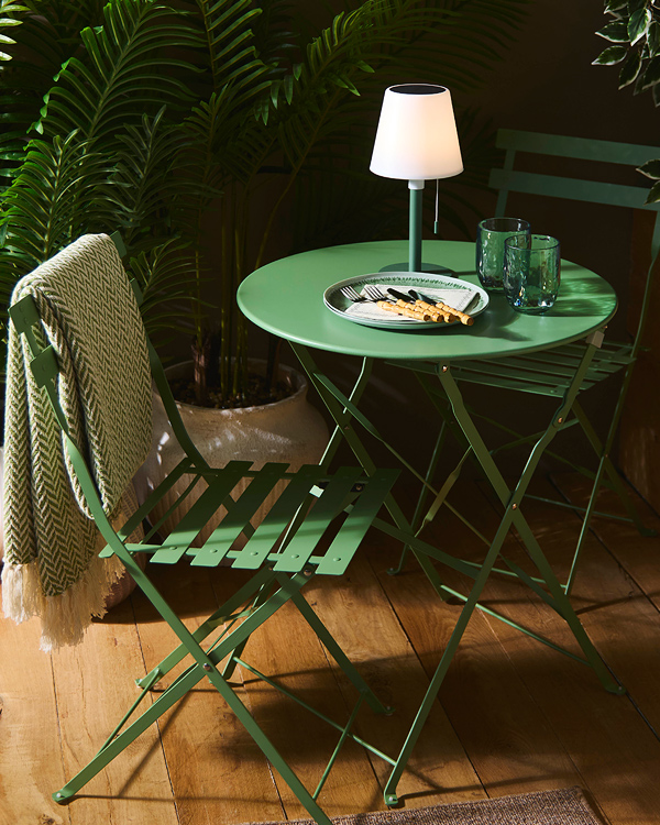 Cozy outdoor setting with green metal table and chairs on wooden floor. White lamp, two glasses, plate with breadsticks, and leafy plants enhance ambiance.