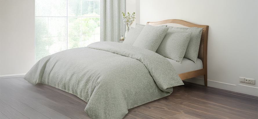 A serene bedroom with a wooden bed and light gray bedding. Large window with curtains lets in soft natural light. A vase with flowers on the side table.
