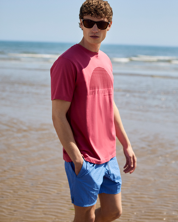 Young man in sunglasses stands on a sunny beach, wearing a pink t-shirt and blue shorts. He has a relaxed, casual demeanor by the ocean.