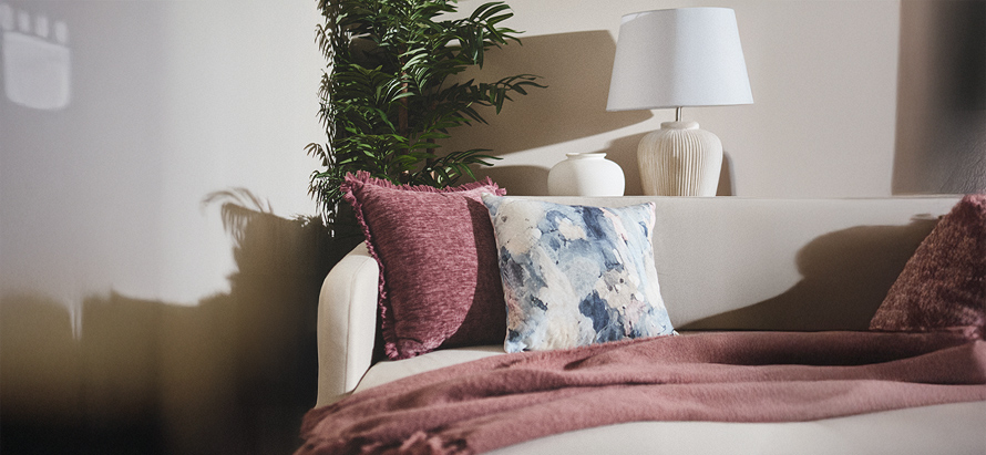 A cozy living room with a cream sofa, adorned with maroon and patterned pillows, a maroon throw blanket, a potted plant, and a white lamp, creating a warm, inviting ambiance.
