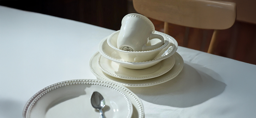 Stacked cream-colored ceramic dishes and a cup on a white table, with soft lighting creating a serene and minimalistic atmosphere. A spoon rests nearby.