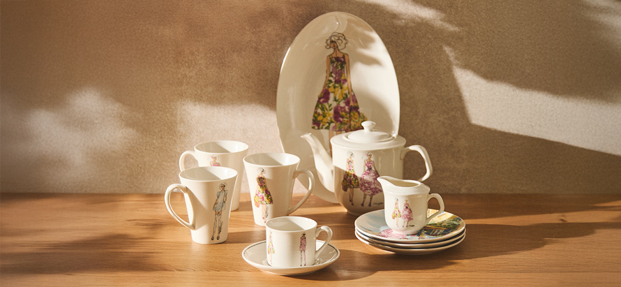 Ceramic tea set on a wooden table with fashion illustrations. Includes cups, saucers, a teapot, and a platter, bathed in warm sunlight. Serene and elegant.