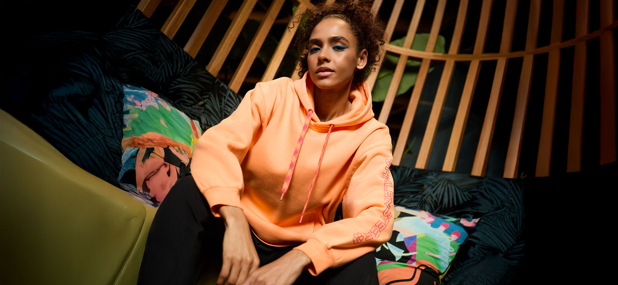 A person sits confidently, wearing a bright orange hoodie, in a modern, artistic space with vibrant, patterned cushions and wooden slats above.