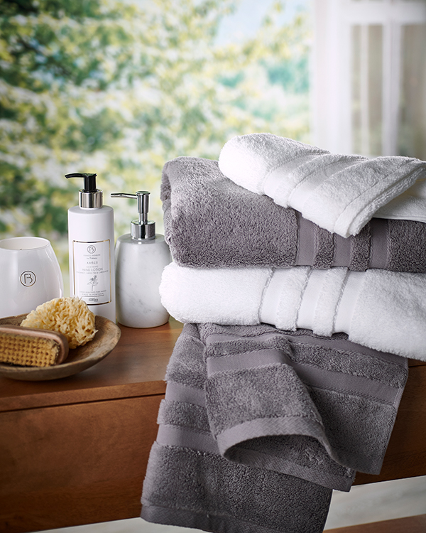 Stack of soft white and gray towels on a wooden shelf with bath essentials like lotion and soap dispensers. Sun-dappled nature view in the background. Cozy and serene.