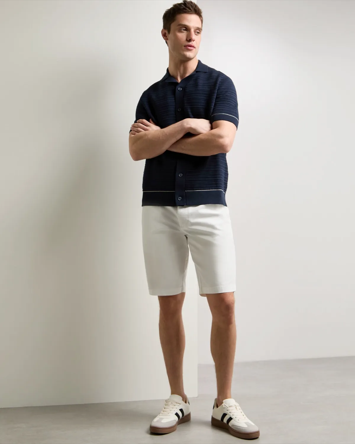 Man in a navy polo and white shorts stands against a light wall, arms crossed, wearing white sneakers with black and brown stripes. Casual and relaxed vibe.