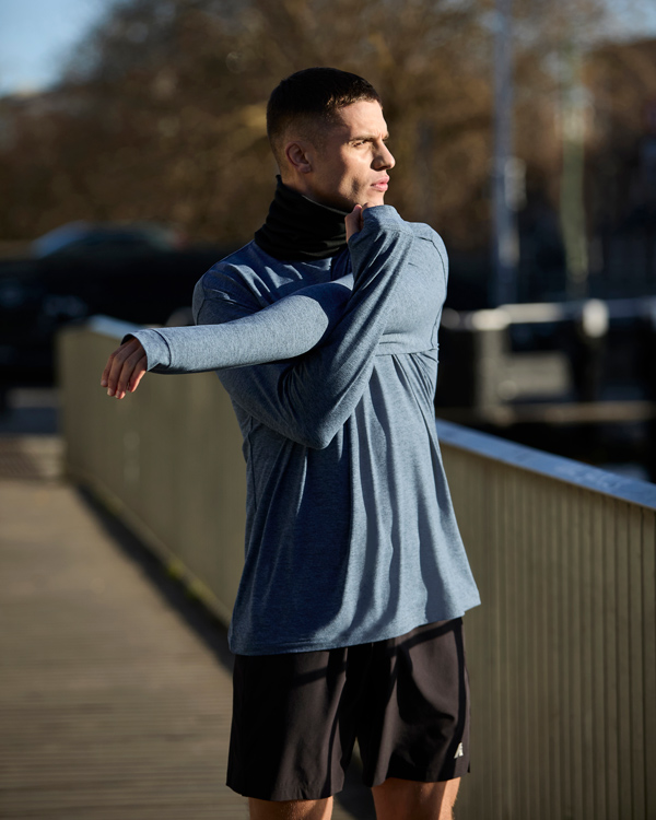 Man in blue long-sleeve shirt stretches on a sunny boardwalk, looking focused and calm. Background features trees and blurred urban elements.
