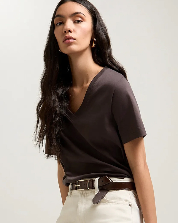 Young woman with long dark hair wearing a brown V-neck T-shirt and white pants with a brown belt. She gazes confidently at the camera against a neutral background.
