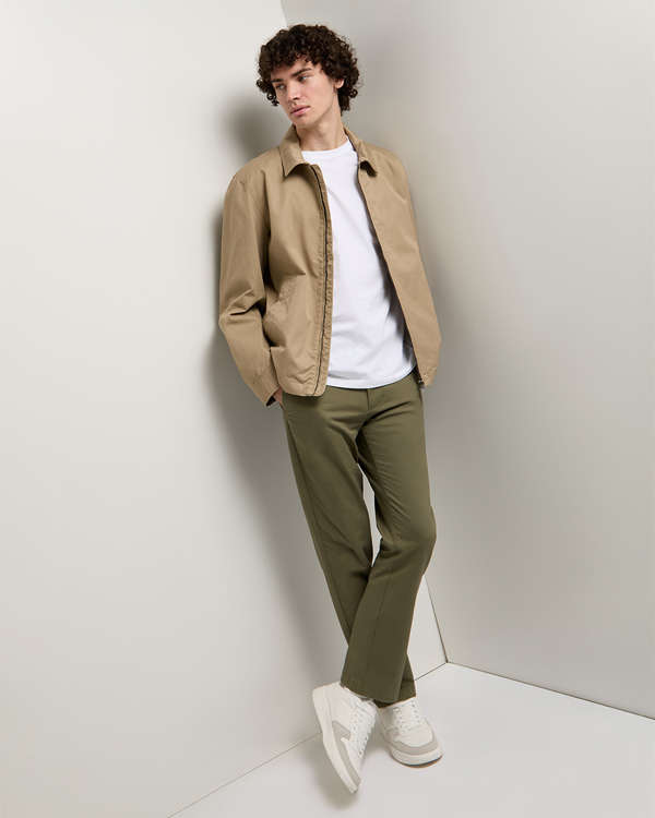 Young man with curly hair leans casually against a white wall. He wears a tan jacket, white t-shirt, olive pants, and white sneakers, conveying a relaxed style.