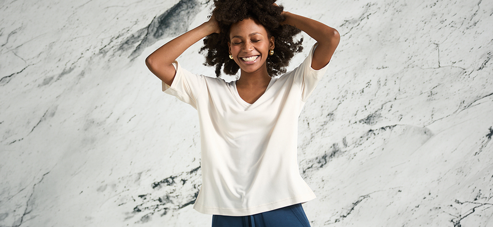 A smiling woman with curly hair wearing a white shirt and blue pants stands against a white marbled background, hands playfully in her hair.