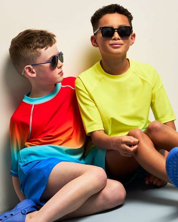 Two boys in vibrant swimwear and sunglasses sit against a wall. One wears a rainbow shirt, the other bright yellow, both exuding a relaxed summer vibe.