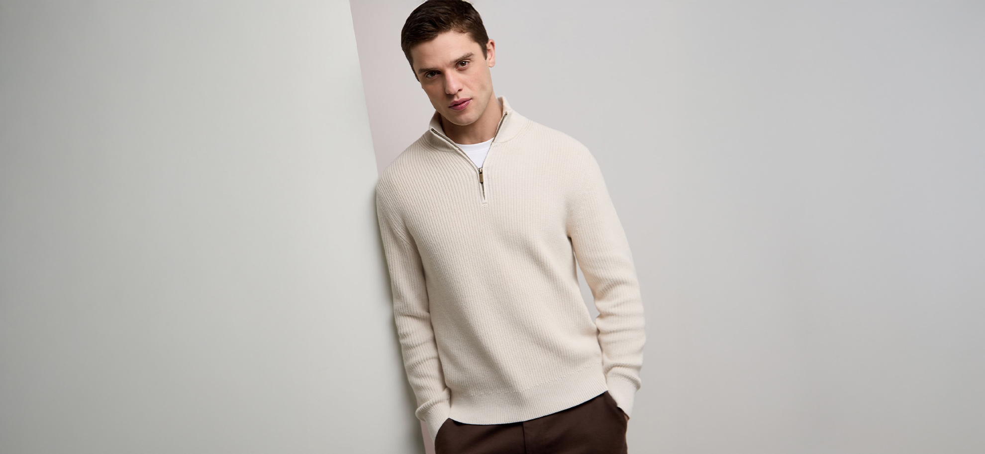 A man leans casually against a light gray wall, wearing a cream-colored zip-up sweater and brown pants. The setting is minimalist and stylish.