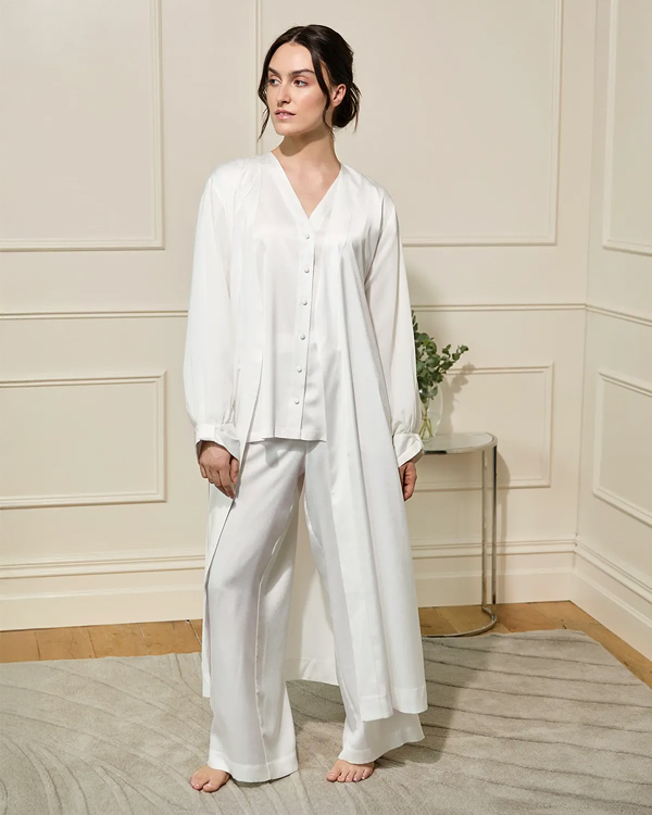 A person in a bright room wearing a flowing white satin pajama set stands on a patterned rug. A small table with a plant is in the background, creating a calm and elegant atmosphere.