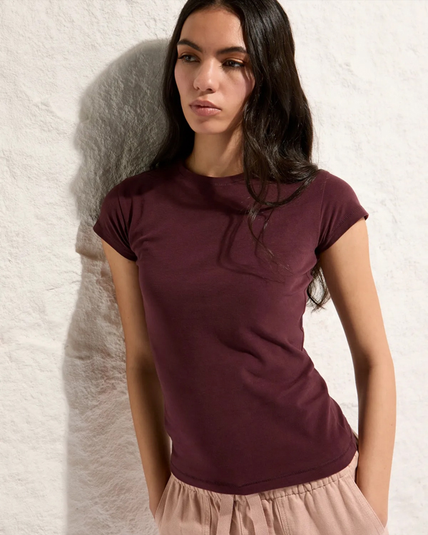 A woman with long dark hair stands against a textured beige wall, wearing a maroon T-shirt and light pink pants. She looks contemplative and calm.