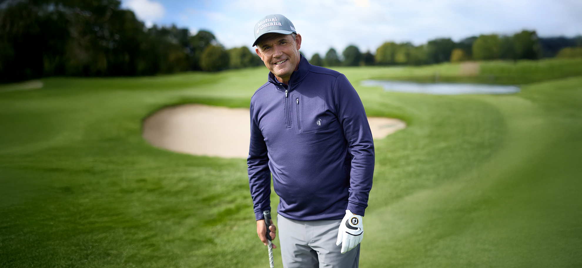 Padraig Harrington in a golf cap and purple jacket smiles on a lush green golf course near a sand bunker. The sky is partly cloudy, creating a cheerful ambiance
