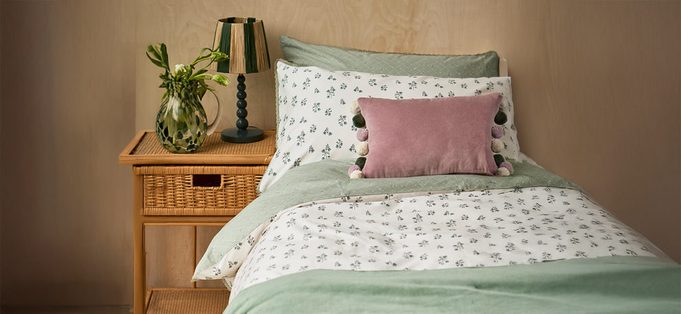 Cozy bedroom scene with a single bed featuring green and white floral bedding and a pink cushion. A wooden nightstand with a vase of flowers and a lamp adds warmth.