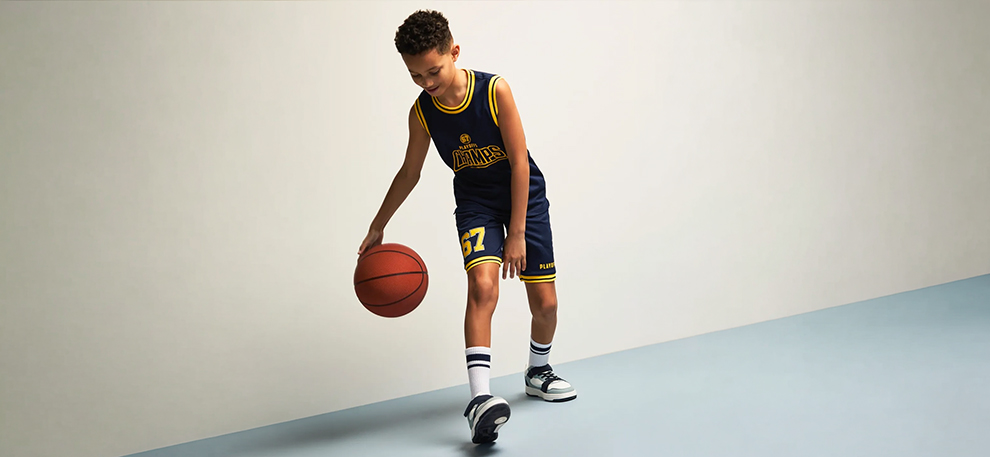 A young boy in a navy and yellow basketball uniform dribbles a basketball on a smooth surface. He looks focused and is wearing sneakers and striped socks.