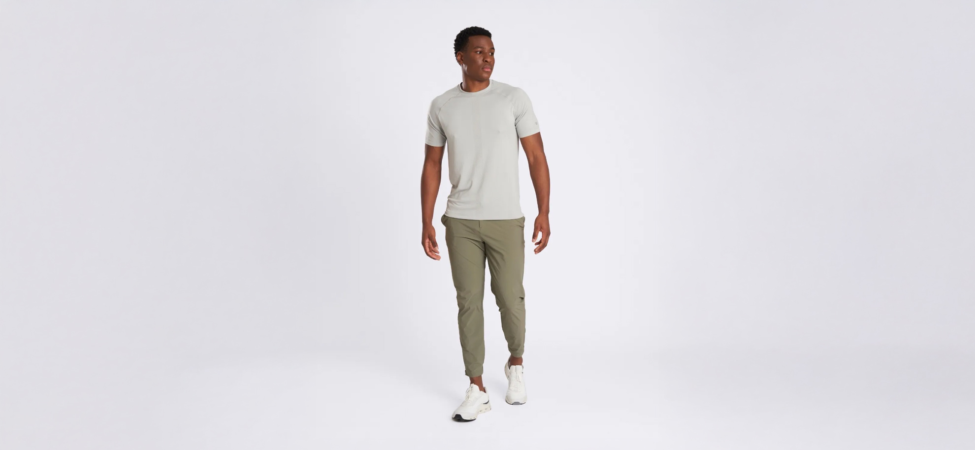 Man in a light gray t-shirt and olive joggers walks confidently against a plain white background. He wears white sneakers, exuding a relaxed, casual style.