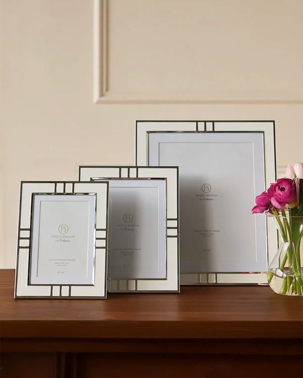Three reflective, silver photo frames of varying sizes with geometric patterns are displayed on a wooden surface. Pink flowers in a vase add a soft touch.
