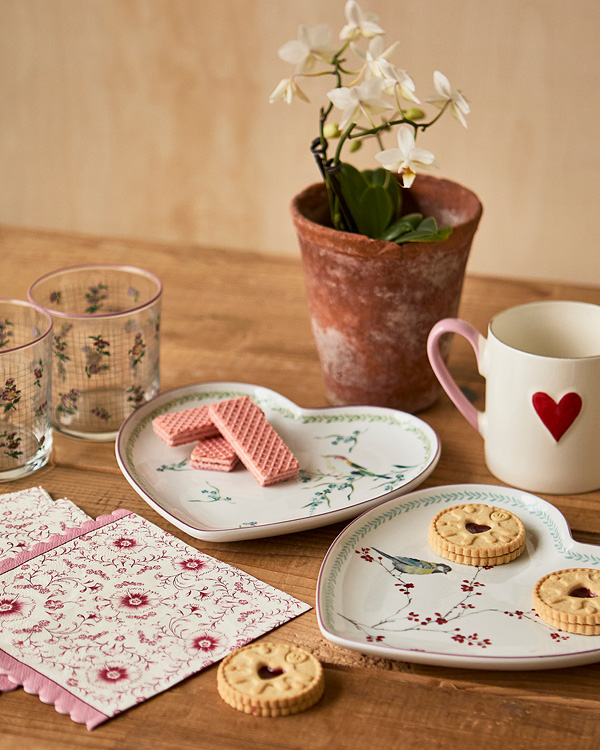 Heart-shaped plates with bird designs hold wafers and cookies on a wooden table. Nearby, two floral glasses, a mug with a heart, and a potted plant.