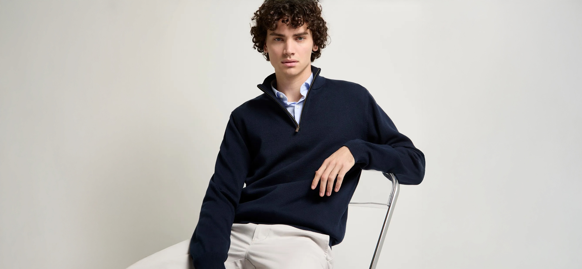 Man wearing a navy fleece, blue shirt and off-white Khaki's, seated on a chair