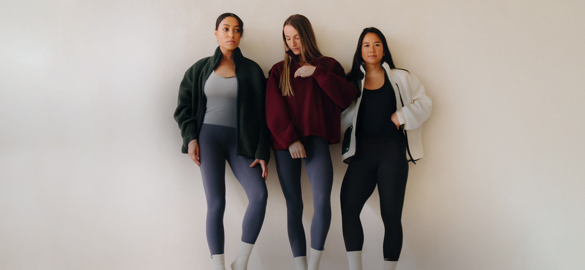 Three women stand confidently against a neutral background, wearing athletic attire: dark leggings, crop tops, and jackets in green, burgundy, and white.