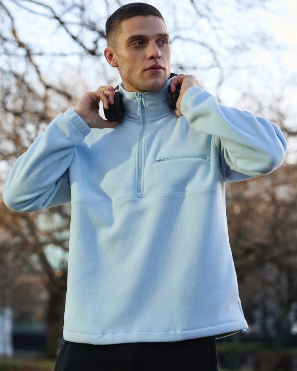 A person stands in a park, wearing a light blue fleece and holding headphones on their shoulders. The background is a blurred view of bare trees.