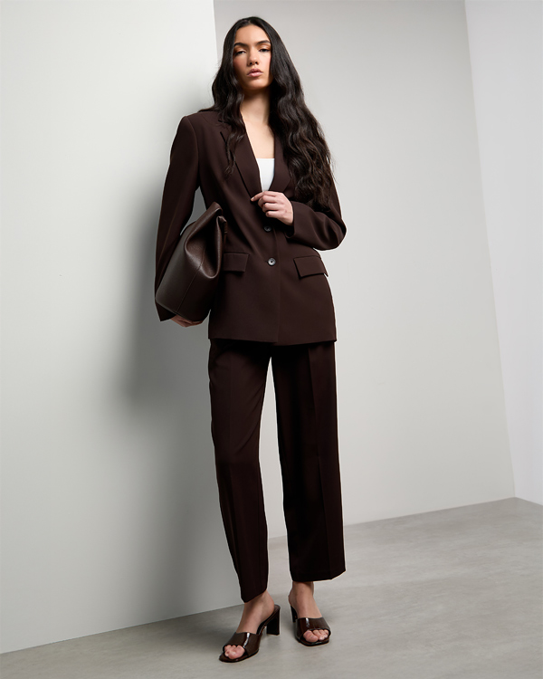 A woman in a sleek brown pantsuit, holding a matching bag, leans stylishly against a light wall. She exudes confidence and modern elegance in heeled sandals.