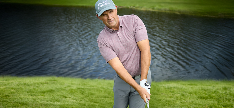Padraig Harrington in a purple polo and gray cap prepares to hit a golf ball near a calm lake. The setting is serene and focused, with lush green grass.