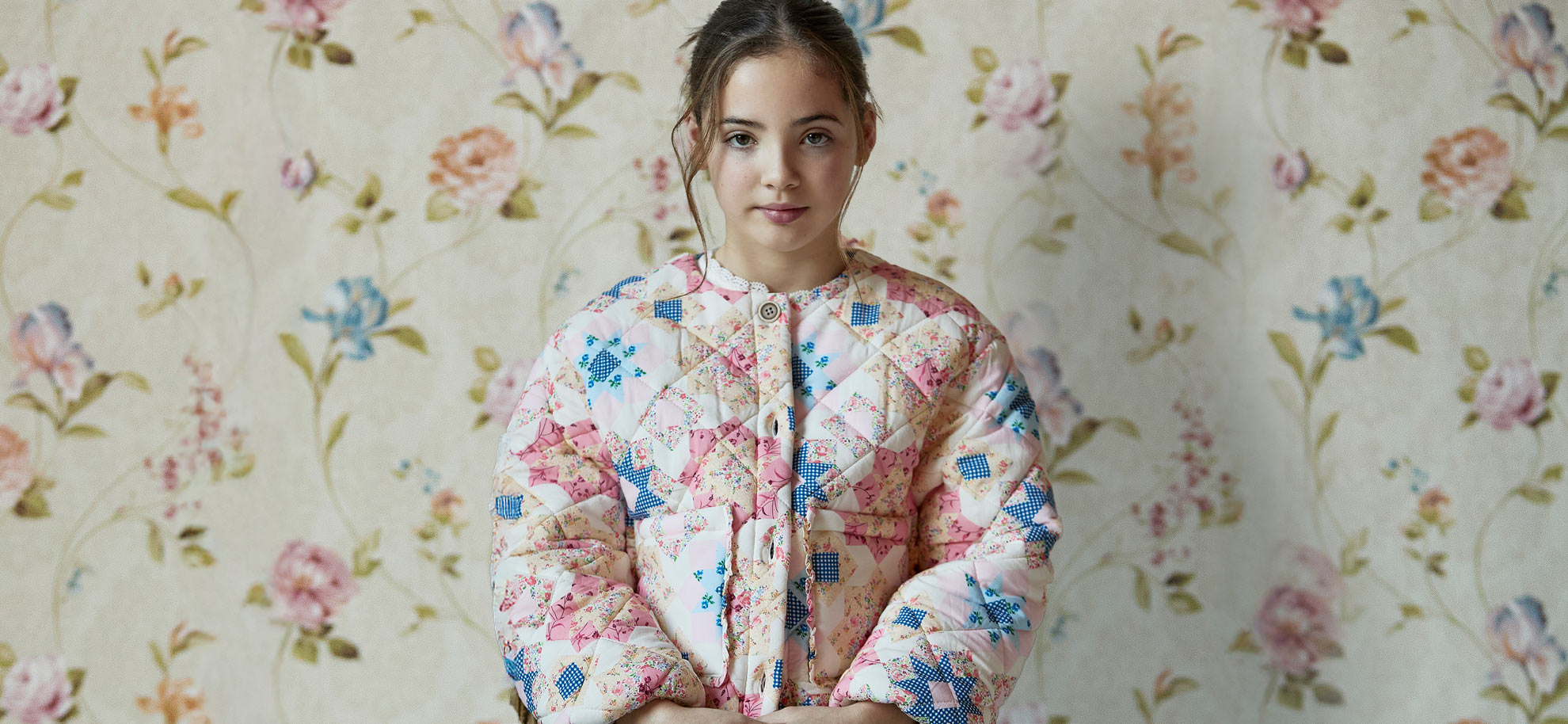 A young girl in a colorful quilted jacket stands against a floral wallpaper background. She has a calm expression, blending with the vintage aesthetic.