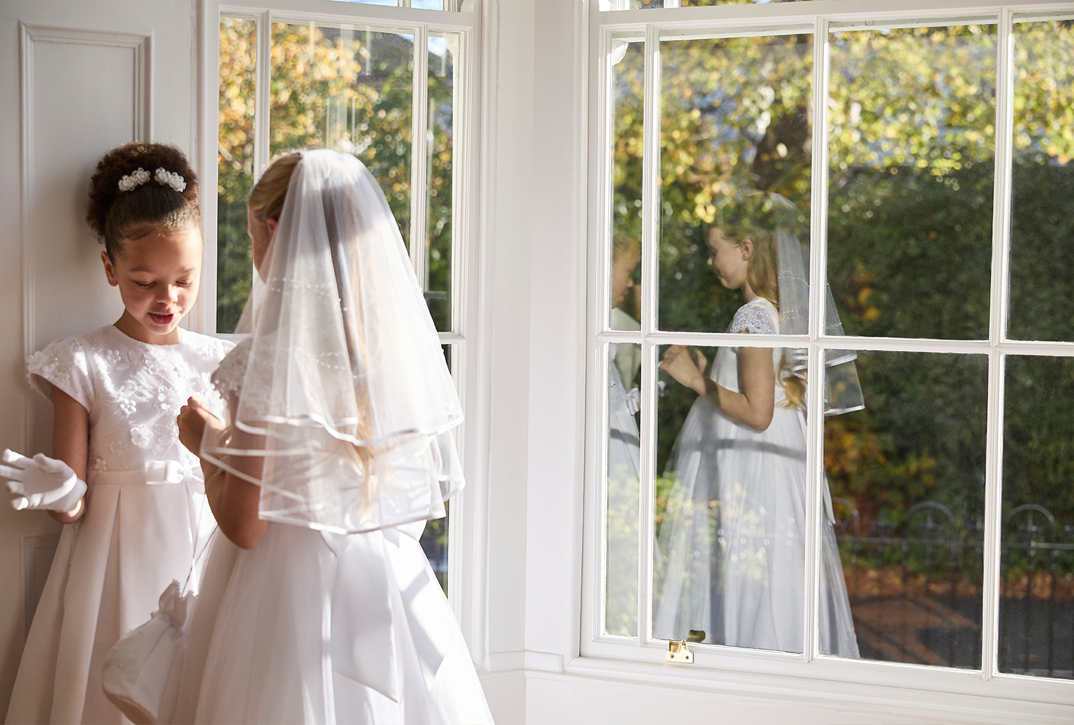 Two young girls in white Paul Costelloe communion dresses stand by a sunny window. One wears a veil, the other has flowers in her hair. Their reflection shows a serene, joyful moment.