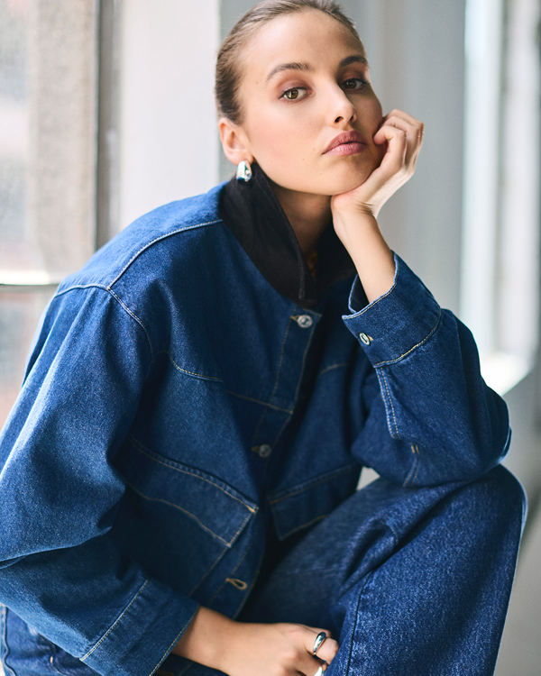 A woman leans her chin on her hand, looking directly forward while wearing a dark denim outfit. The medium-blue denim jacket features a collarless neckline with a buttoned front placket and is layered over a high-collared black top. She pairs it with matching blue denim jeans, accessorizing with bold, sculptural silver earrings and a thick silver dome ring on her right hand. 