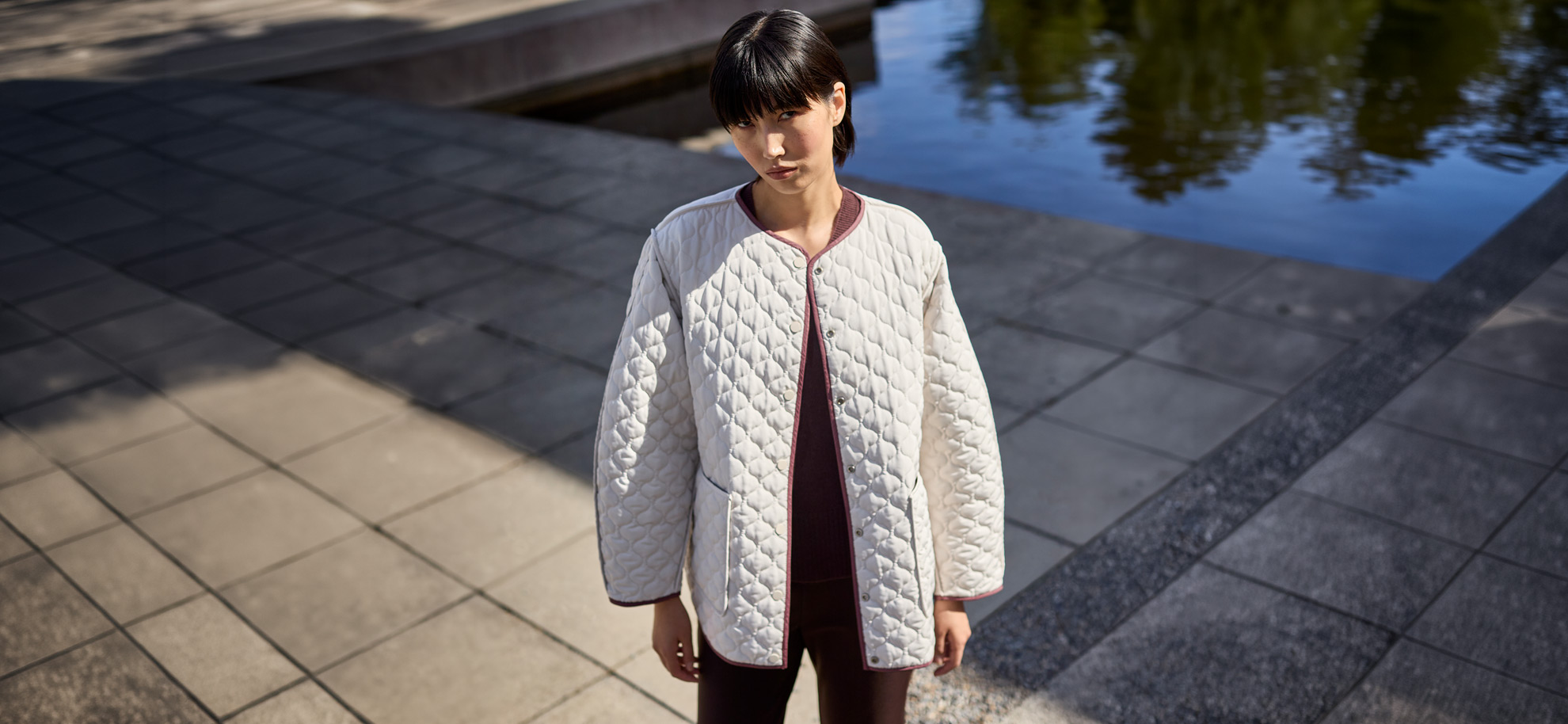 A woman with short black hair and bangs stands outdoors, looking directly forward while modeling a collarless, quilted cream jacket with an open front secured by metal snap fasteners. The mid-length outerwear features a distinct diamond-quilt pattern, wide sleeves, and contrasting dark piping along the edges, worn over a dark burgundy top and matching pants. She is positioned on a grey stone tiled pathway next to a calm body of water that reflects the surrounding trees and shadows under bright sunlight.