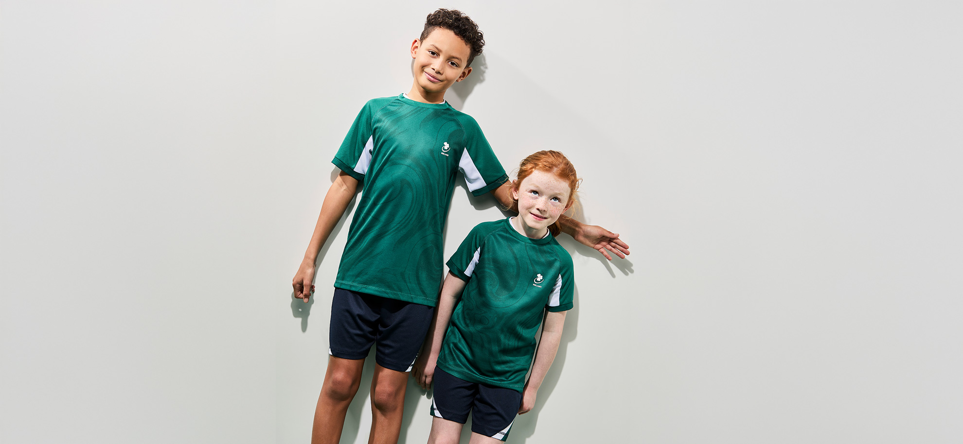 Two children wearing matching green sports jerseys and navy shorts stand against a light background. They smile, exuding a playful and cheerful vibe.