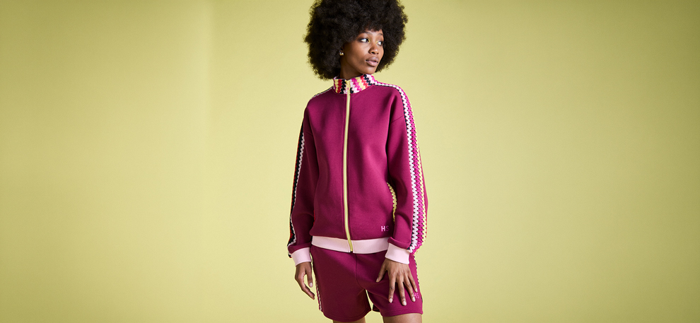 A woman in a matching pink tracksuit poses against a light green background. She looks to the side with a confident expression, hands relaxed.