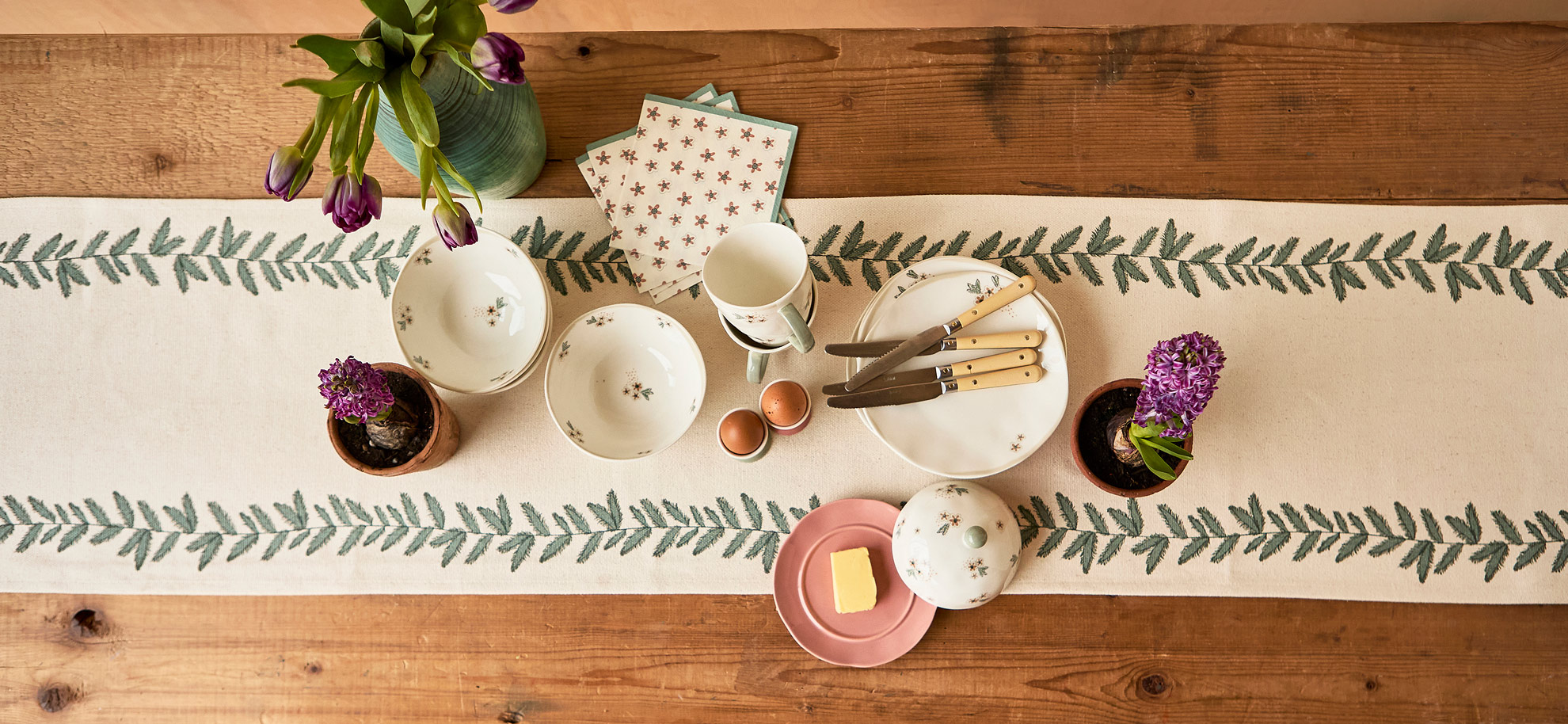 A rustic table setting with a white runner adorned with green leaf patterns. Plates, mugs, and cutlery are arranged neatly alongside purple flowers, eggs, and butter.