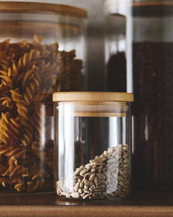 A small, clear glass storage jar with a light-brown bamboo lid sits in the foreground, partially filled with sunflower seeds resting at a diagonal angle. In the softly blurred background, two larger matching glass canisters line the shelf, with one on the left tightly packed with spiral fusilli pasta and the other on the right containing dark grains. The composition uses a warm, rustic color palette and shallow depth of field to showcase an organized and aesthetic kitchen pantry setup.