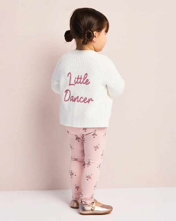 A toddler stands against a light pink background, wearing a white sweater with "Little Dancer" in pink, floral leggings, and gold shoes. The mood is playful.