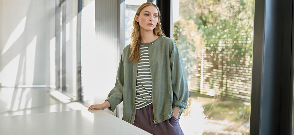Young woman in a casual green jacket and striped shirt stands pensively by a window. Soft light filters in, creating a calm, contemplative mood.