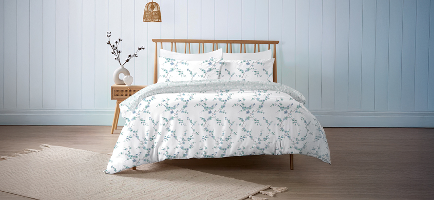 A cozy bedroom features a light wooden bed with floral-patterned bedding, a bedside table with elegant decor, a woven lamp, and a serene ambiance.