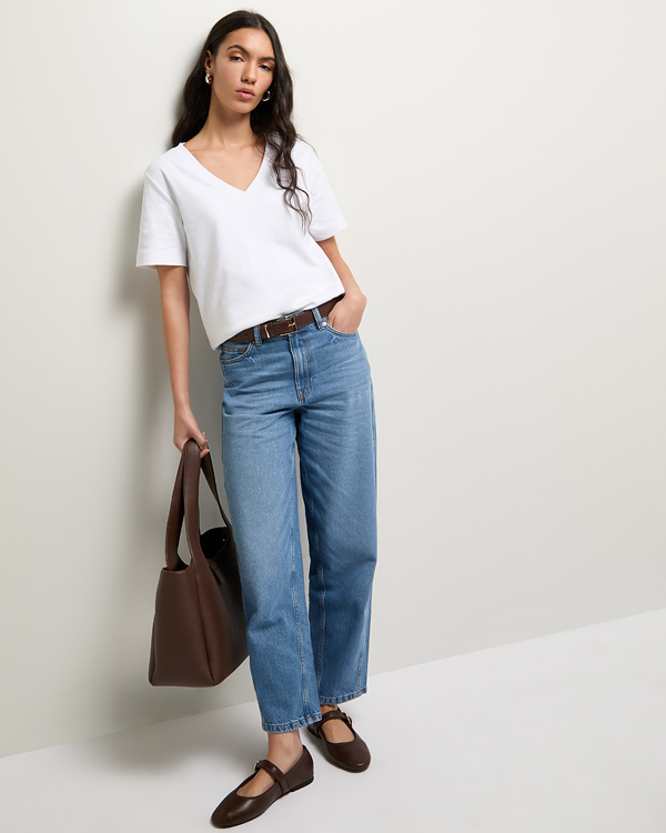 Woman in a casual outfit leans against a wall. She wears a white V-neck T-shirt, blue jeans, brown belt, and shoes, holding a large brown bag. Relaxed and stylish tone.