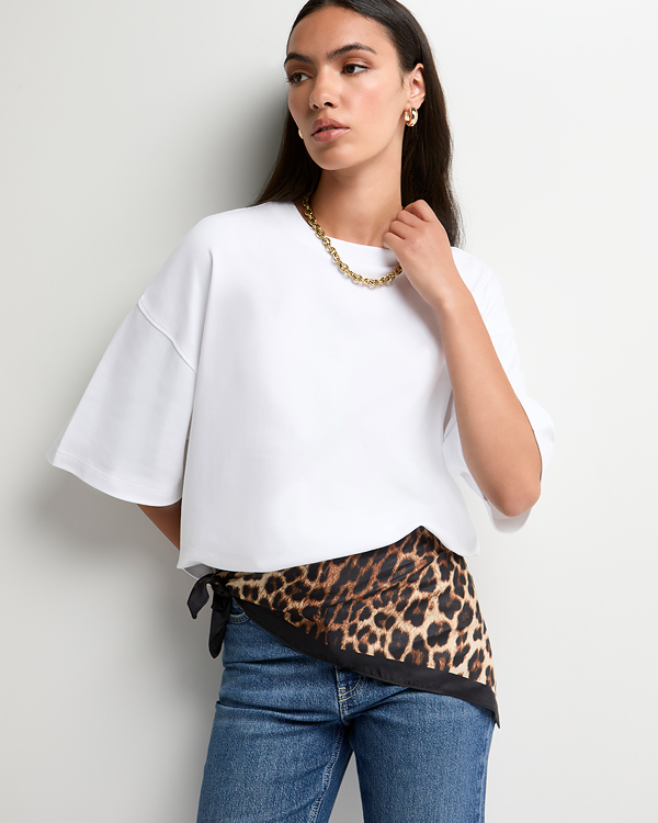 A woman poses against a white background wearing an oversized white top with dramatic, elbow-length drop sleeves. A satin-like leopard-print fabric with a black border peaks out asymmetrically below the hem, knotted at her hip. She pairs the top with medium-wash blue jeans and details the look with a chunky gold link chain necklace and matching thick hoop earrings. Her long dark hair is neatly tucked back, and she looks thoughtfully off-camera to the left.