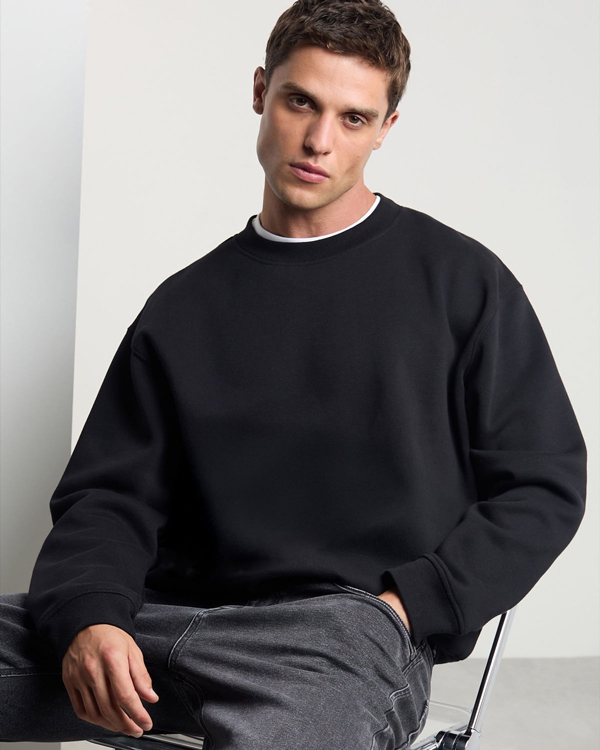 Young man wearing a black sweatshirt and gray jeans sits on a chair against a plain background, with a contemplative expression. Casual and minimalistic.