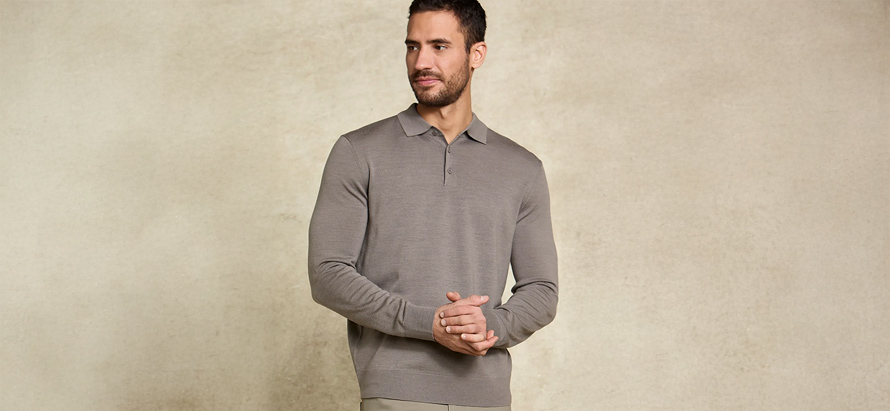 A man in a neutral-colored, long-sleeve polo shirt stands against a plain beige background, looking to the side with a calm expression and clasped hands.