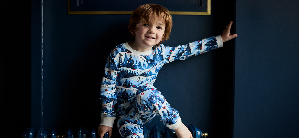 A young boy with reddish hair leaning against a dark blue wall, wearing blue long-sleeve pajamas featuring a winter forest and skiing penguin print.