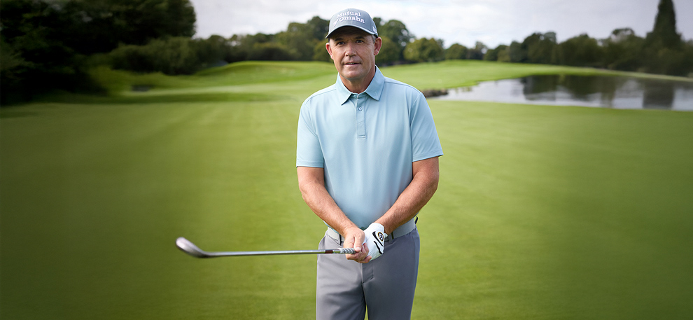 Padraig Harrington in a light blue polo and cap holding a golf club stands on a lush green golf course, with trees and a pond in the background, conveying calm focus.
