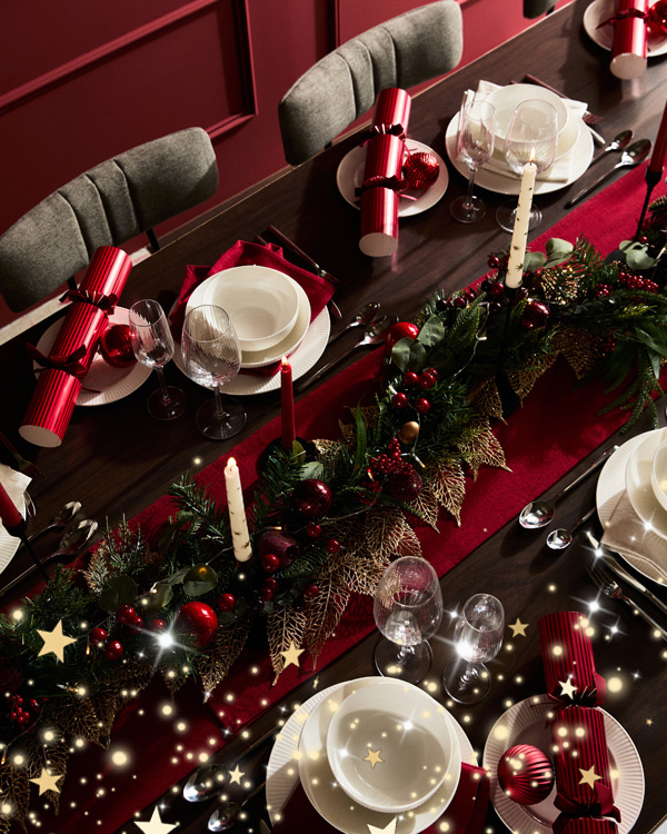 Festive table setting with red and white plates, glasses, crackers, and candles on a dark wood table. Greenery and gold accents create a holiday atmosphere.