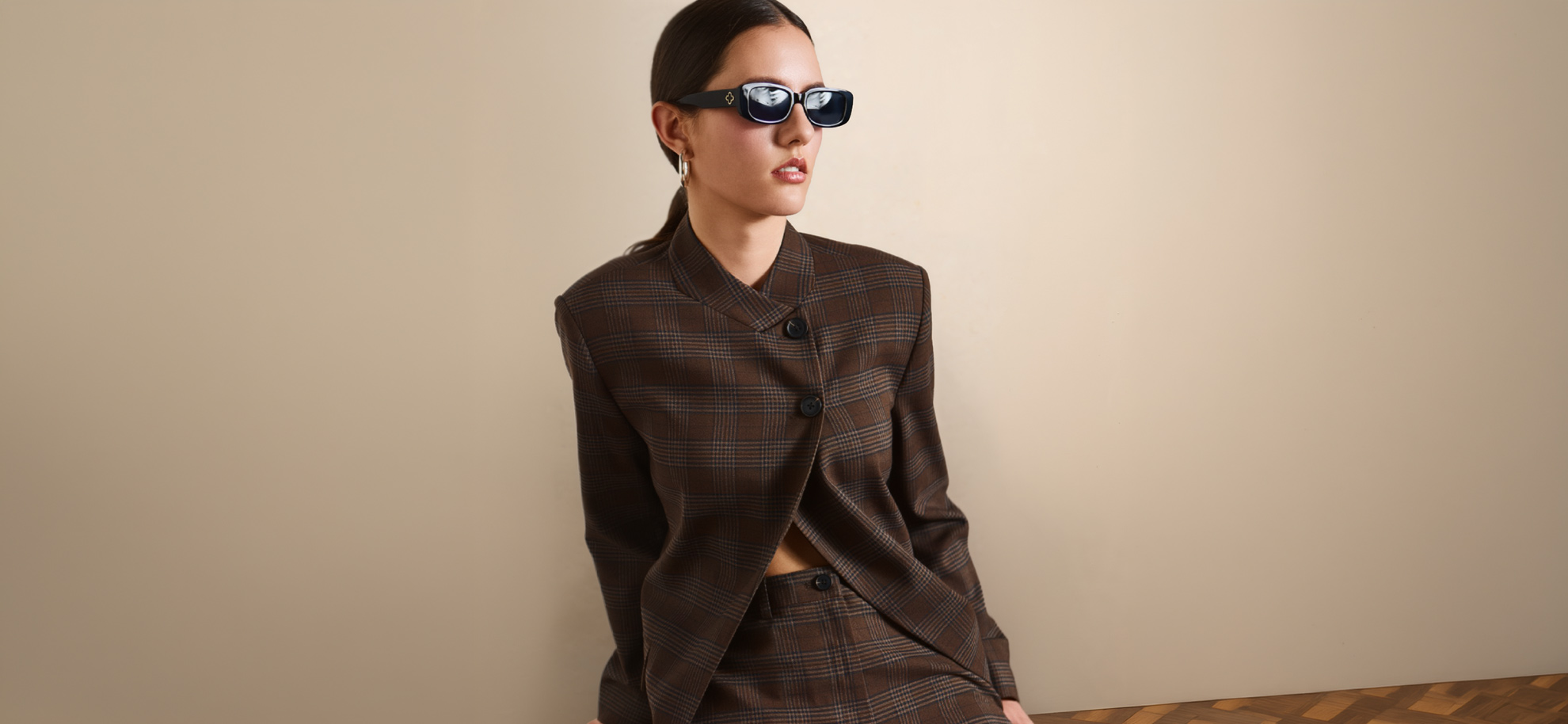 A woman with dark hair pulled back poses in a structured, brown plaid blazer and matching trousers against a solid beige wall. The blazer features an asymmetric button closure and a mandarin collar. She wears rectangular black sunglasses and simple silver hoop earrings, looking slightly to her right.