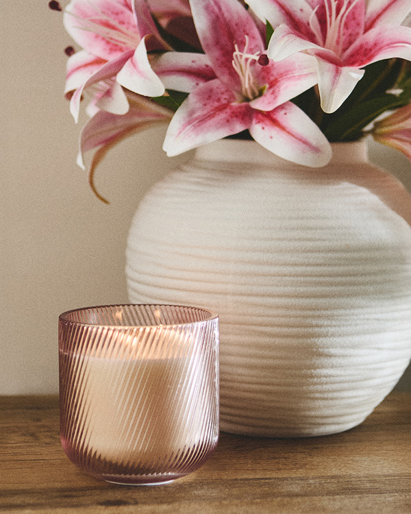 A soft pink candle in a striped glass holder sits on a wooden table beside a white textured vase with blooming pink lilies. The scene is serene and elegant.