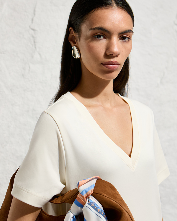 A woman with long, straight dark hair parted down the middle poses against a textured white brick wall. She wears a minimalist cream-colored, short-sleeved top with a V-neckline. She is accessorized with large, sculptural silver statement earrings. A portion of a brown suede bag is visible in the lower foreground, featuring a blue, white, and orange patterned satin scarf tied around its handle. She looks directly forward with a calm, neutral expression.