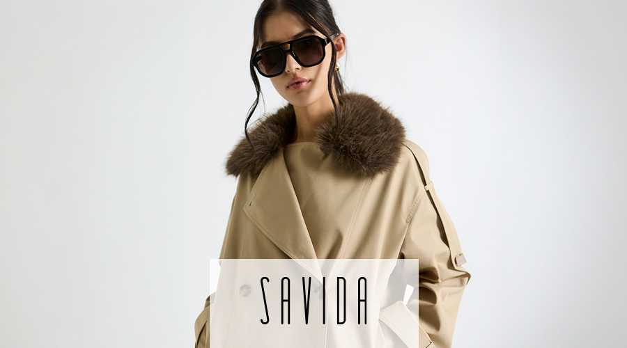 A young woman with dark hair tied back poses against a plain white background, wearing large, dark aviator-style sunglasses. She is dressed in a beige trench coat featuring a prominent, fluffy brown faux-fur collar that wraps around her neck. Over the lower portion of the image, the brand name "SAVIDA" is written in a stylized, tall black font.